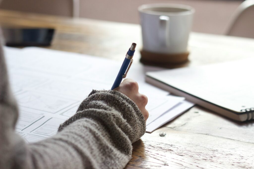 Person writing on paper with a pen, cozy sweater sleeve visible.